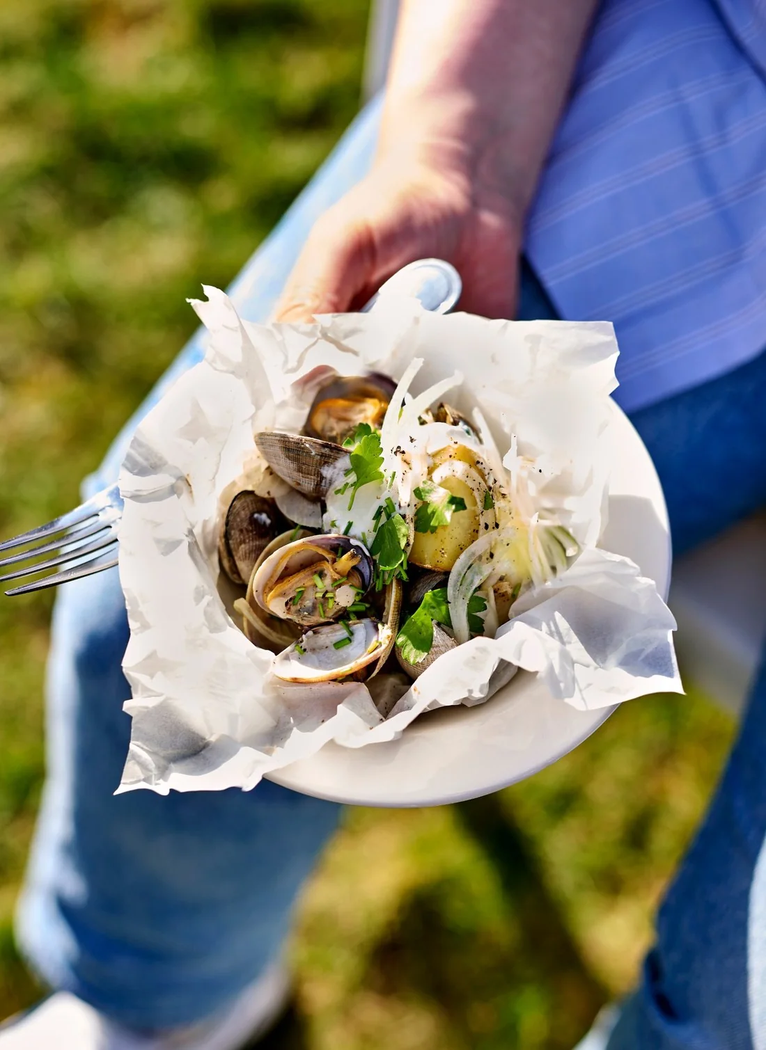 Shot on set: someone in jeans on the grass holds a bowl of clams and potatoes in baking paper